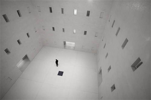 A lone person standing in a vast, empty concrete room with many small windows